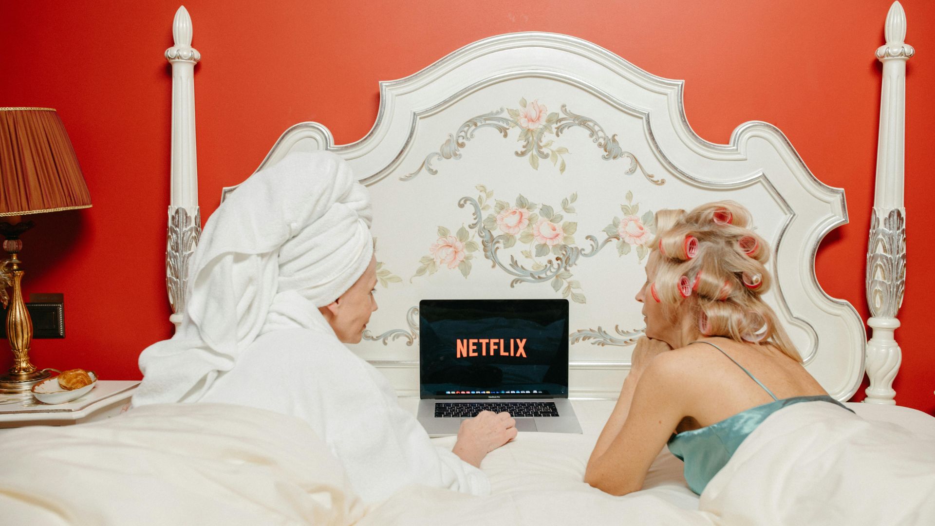 Serien Highlights 2025: Zwei Frauen liegen auf dem Bauch in einem Bett und schauen auf einen Laptop mit dem Netflix Startbildschirm. Die linke Frau trägt Bademantel und hat ein Handtuch um ihre Haare gewickelt. Die rechte Frau hat viele Lockenwickler in ihren blonden Haaren. Das Bett steht vor einer roten Wand.