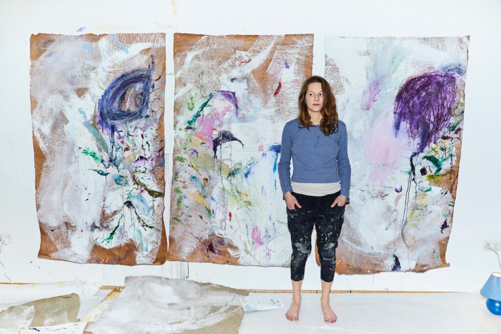 Ina Fasching steht barfuß vor zwei großformatigen Gemälden mit expressiv-abstrakter Malerei in ihrem Atelier.