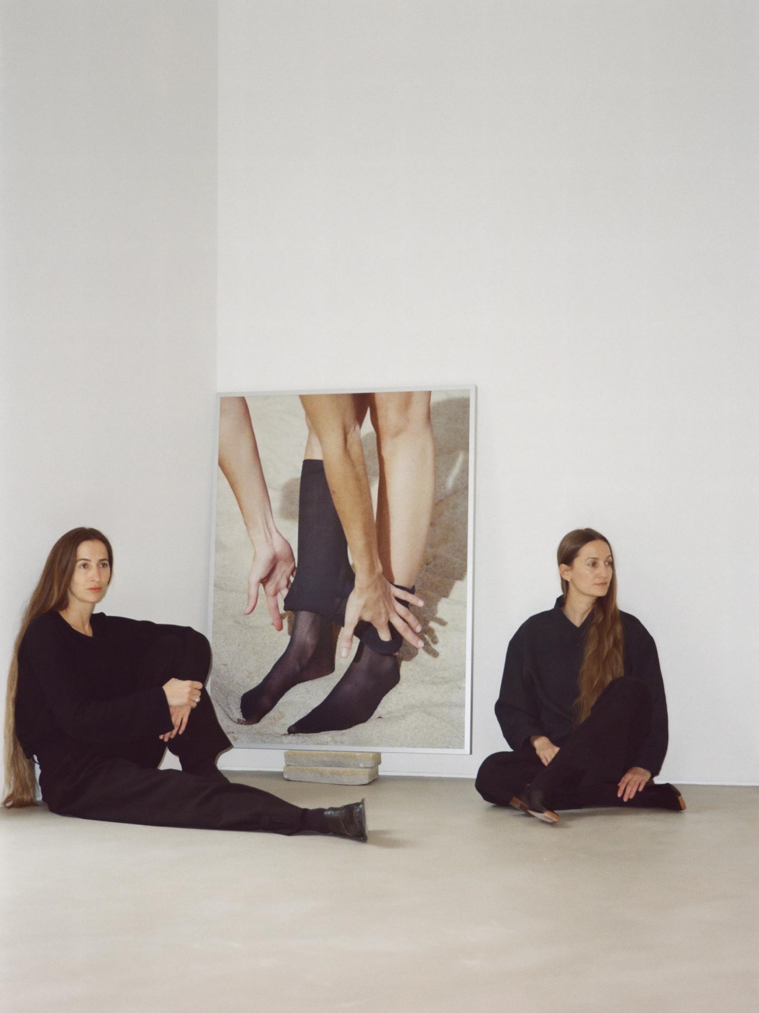 Anna und Maria Ritsch sitzen neben einem ihrer Werke – ein intimes Fotomotiv mit femininer Körperdarstellung, aufgenommen in minimalistischer Galerieatmosphäre.