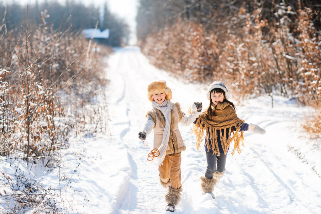 Zwei Kinder laufen glücklich auf einem schneeigen Weg im Wald mit Haus im Hintergrund und blätterarmen Büschen an den Seiten.