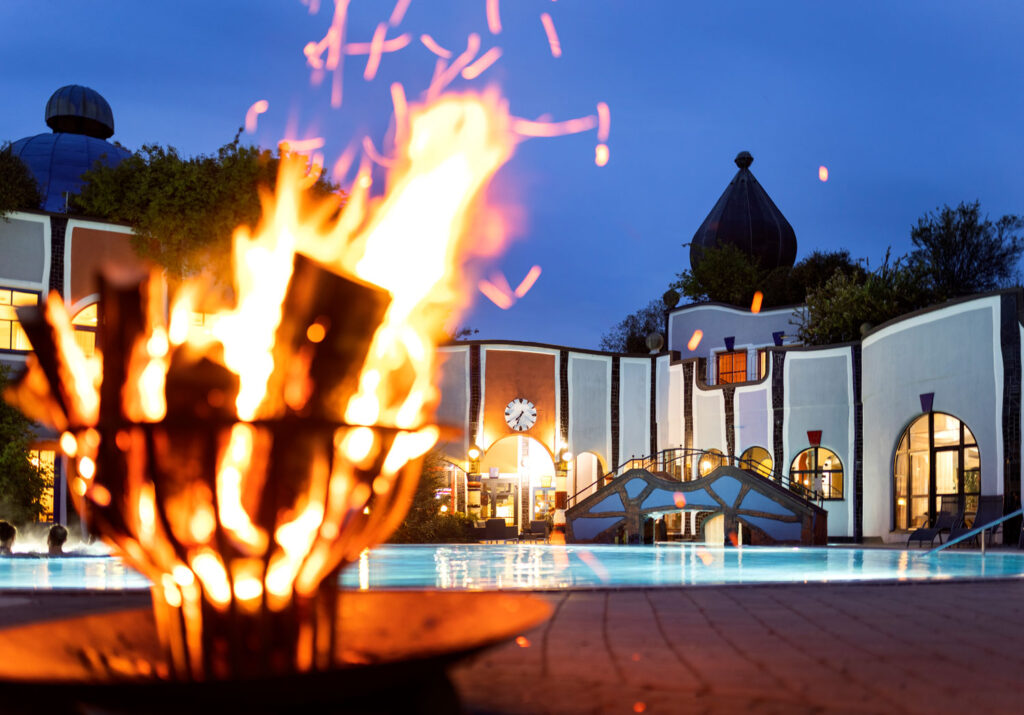 Rogner Bad Blumau mit Thermenpool von außen und einem Feuer, das unscharf im Vordergrund brennt.