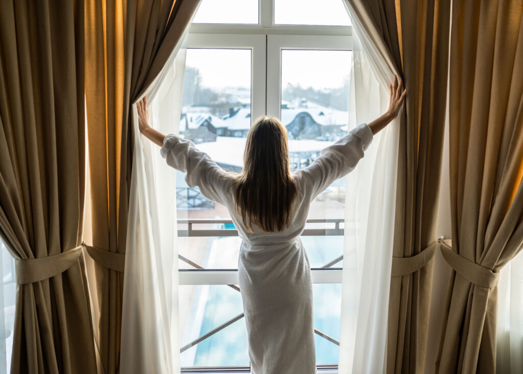 Weekend Retreats : Frau im Bademantel öffnet Vorhang mit winterlicher Aussicht in einem Hotelzimmer.