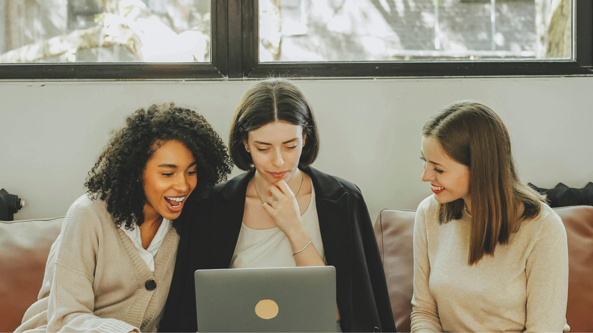 Drei Frauen sitzen gemeinsam auf einem Sofa. Die mittlere schaut auf ihren Laptop, die anderen lächeln.